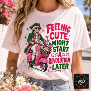 May include: A white t-shirt featuring a graphic of a historical figure in a green coat and heart-shaped sunglasses riding a pink scooter. The text reads: Feeling CUTE, MIGHT START A REVOLUTION LATER. A small American flag and flowers are on the scooter.