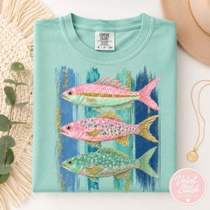 Pode incluir: Uma t-shirt verde-menta claro com um gr&aacute;fico de tr&ecirc;s peixes empilhados verticalmente. Os peixes s&atilde;o representados em tons de rosa, menta e glitter dourado. O fundo do gr&aacute;fico consiste em pinceladas verticais azuis e menta com detalhes.