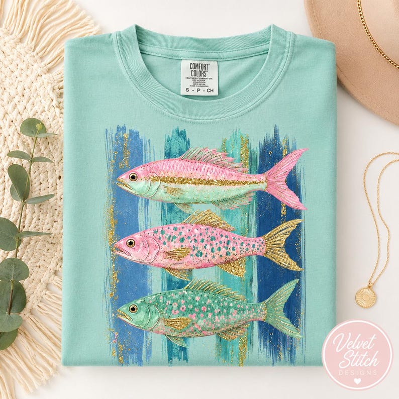 Pode incluir: Uma t-shirt verde-menta claro com um gr&aacute;fico de tr&ecirc;s peixes empilhados verticalmente. Os peixes s&atilde;o representados em tons de rosa, menta e glitter dourado. O fundo do gr&aacute;fico consiste em pinceladas verticais azuis e menta com detalhes.