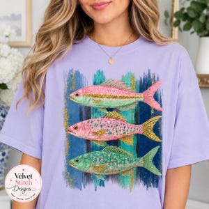Pode incluir: Uma t-shirt roxo-claro com uma estampa gr&aacute;fica de tr&ecirc;s peixes empilhados verticalmente. Os peixes s&atilde;o representados em tons de rosa, verde e dourado com detalhes brilhantes, sobre um fundo de pinceladas verticais azuis e verdes.