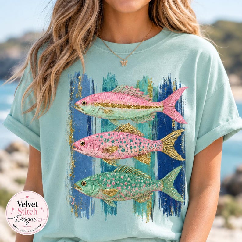 Pode incluir: Uma t-shirt verde menta claro com um gr&aacute;fico de tr&ecirc;s peixes empilhados. O peixe superior &eacute; rosa e menta, o do meio &eacute; rosa com glitter dourado e o inferior &eacute; verde menta com glitter dourado, sobre tra&ccedil;os de pincel azuis verticais.
