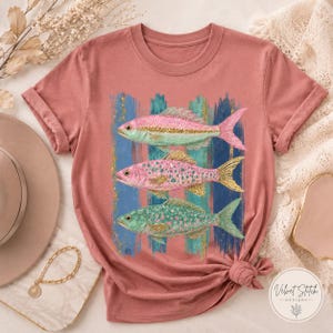 Pode incluir: Uma t-shirt de manga curta em rosa seco com uma impress&atilde;o gr&aacute;fica de tr&ecirc;s peixes sobrepostos. Os peixes s&atilde;o representados em tons de rosa, azul-petr&oacute;leo e dourado com detalhes brilhantes sobre um fundo de pinceladas verticais azuis e esverdeadas.