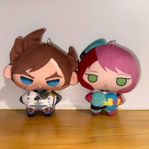 Puede incluir: Dos muñecos de peluche con rasgos de estilo anime. Uno tiene el pelo castaño y los ojos azules, el otro tiene el pelo rosa y los ojos verdes. Ambos muñecos tienen trajes detallados y están diseñados con un lazo para llavero.
