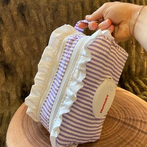 Peut inclure: Une pochette en tissu rectangulaire avec un motif à rayures violettes et blanches et des bordures à volants blancs. La pochette comprend un couvercle distributeur à clapet en plastique blanc avec le texte rouge HUGGIES imprimé dessus.