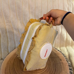 May include: A mustard yellow and white striped diaper wipes case with a white zip and ruffled trim. The case has a handle and a white oval sticker with the word "Huggies" in red. The case is on a wooden surface.