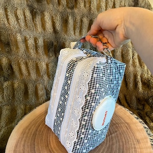 May include: A black and white gingham patterned wipe dispenser with white eyelet lace trim and a white Huggies label. The dispenser has a handle and a silver-coloured clasp.