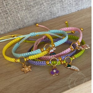 May include: A collection of colorful macrame bracelets with gold-colored accents and charms. The bracelets are in shades of yellow, blue, pink, purple, and green. Charms include a flower, a bunny, a flower with a purple center, and a leaf.