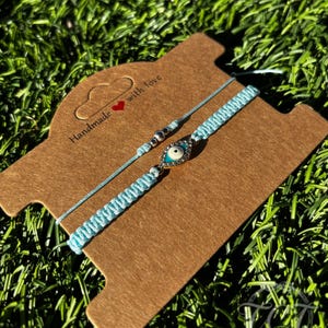 May include: Two light blue handmade bracelets with an evil eye charm and silver beads are displayed on a brown card. The card has the text "Handmade with love" and a red heart. The background is green grass.