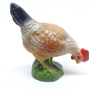 May include: A vintage-style cast metal figurine of a chicken in a pecking position. The bird features a brown and cream-colored body, a dark blue tail, and a bright red comb. It stands on a textured green base representing grass.