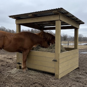 Op de afbeelding: Een lichtbruine houten paardenvoederbak met een dak van golfplaten. De structuur heeft een vierkant frame met horizontale houten planken en zwarte metalen scharnieren. Twee kastanjebruine paarden eten hooi uit het midden van de bak.