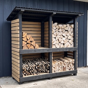 May include: A two-level outdoor firewood storage rack featuring a black painted wooden frame and natural light-colored horizontal wooden slats. The structure includes a corrugated black metal roof and is filled with various sizes of stacked firewood.