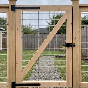 K&ouml;nnte beinhalten: Ein Holztor mit einem hellbraunen Zedernholzrahmen, einer diagonalen St&uuml;tzstrebe und einem silbernen Drahtgittereinsatz. Das Tor ist mit zwei schwarzen Metallscharnieren auf der linken Seite und einem schwarzen Metall-Schieberiegel ausgestattet.