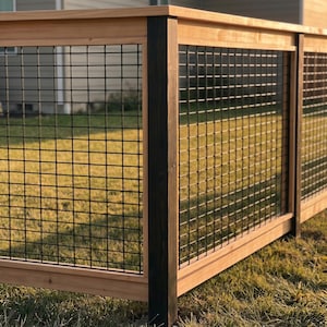 May include: A wooden fence section featuring a natural light brown horizontal frame and a dark stained vertical post. The structure incorporates a black metal wire grid panel with a square mesh pattern, creating a clean and structured barrier design.