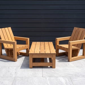 May include: A set of outdoor furniture consisting of two wooden armchairs and a matching rectangular coffee table. Each chair features a slatted backrest and seat with thick, square-profile armrests and a sturdy base. The table has a slatted top.