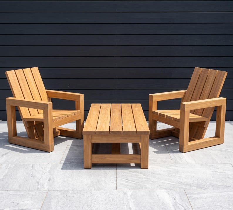 May include: A set of outdoor furniture consisting of two wooden armchairs and a matching rectangular coffee table. Each chair features a slatted backrest and seat with thick, square-profile armrests and a sturdy base. The table has a slatted top.