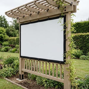 Op de afbeelding: Een houten buitenscherm met een wit projectieoppervlak. Het scherm is zwart omlijst en gemonteerd op een houten constructie met een pergola-stijl dak. Groene wijnranken klimmen op de palen in een tuin.