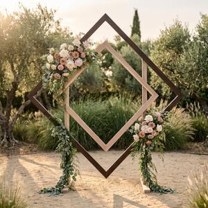 May include: A large, double-layered diamond-shaped wooden wedding arch featuring a dark brown outer frame and a lighter wood inner frame. The structure is decorated with two floral arrangements of pink, cream, and peach roses with green eucalyptus.