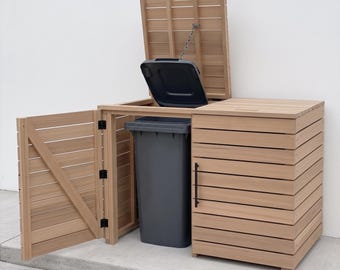 May include: A light brown wooden trash bin enclosure featuring horizontal slats, a hinged door with a black handle, and a top lid attached to a chain. The structure houses a tall, dark gray plastic waste bin with its own lid open.