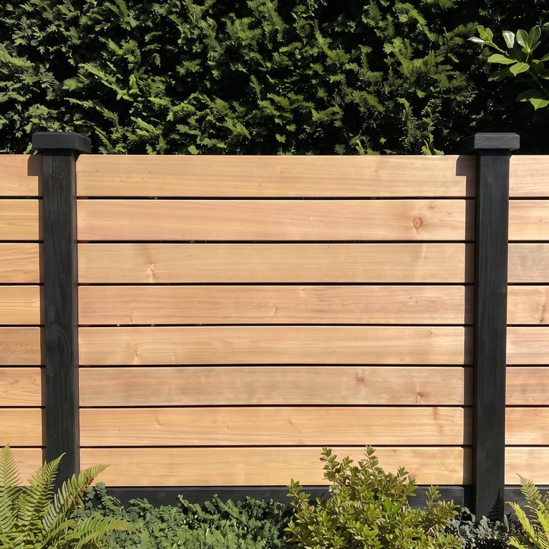 May include: A horizontal wood slat fence featuring natural light brown cedar planks arranged in a stacked pattern. The fence is supported by two vertical black painted wooden posts with square caps, creating a contrast between the wood tones.