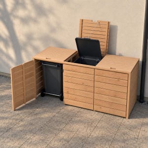 May include: A light brown wooden trash bin enclosure with three compartments. The left door is open, revealing a dark gray wheeled trash bin. The middle compartment lid is open, showing the interior, while the right lid remains closed.