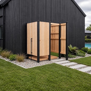 May include: An outdoor shower enclosure featuring vertical light-toned wooden slats, a matching wooden floor base, and a black metal frame. The structure is positioned against a black exterior wall, with a black pipe visible on the side panel.