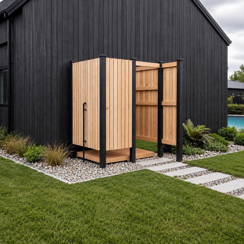 May include: An outdoor shower enclosure featuring vertical light-toned wooden slats, a matching wooden floor base, and a black metal frame. The structure is positioned against a black exterior wall, with a black pipe visible on the side panel.