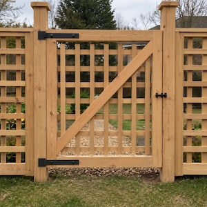 K&ouml;nnte beinhalten: Ein hellbraunes Holztorgartentor mit quadratischem Gittermuster und einer diagonalen St&uuml;tzstrebe. Das Tor ist zwischen zwei dicken Holzpfosten montiert und verf&uuml;gt &uuml;ber schwarze Metallscharniere sowie einen schwarzen Riegel.