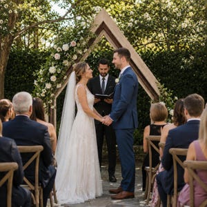 May include: A wedding ceremony featuring a couple standing under a triangular wooden arch decorated with white and pink flowers. The individual on the left wears a white lace gown and veil, while the individual on the right wears a blue suit.