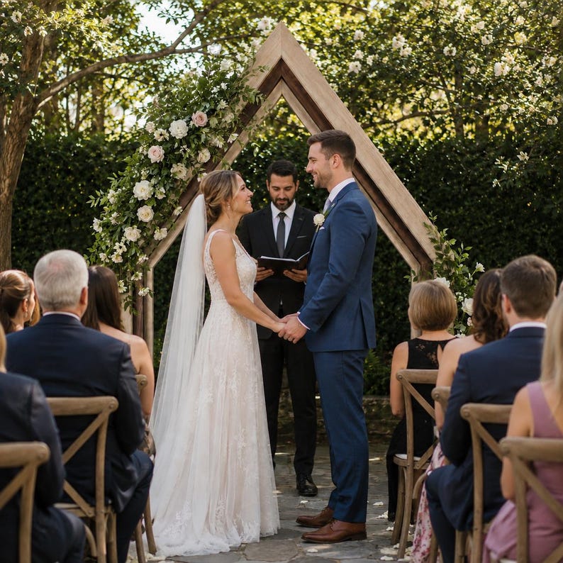 May include: A wedding ceremony featuring a couple standing under a triangular wooden arch decorated with white and pink flowers. The individual on the left wears a white lace gown and veil, while the individual on the right wears a blue suit.