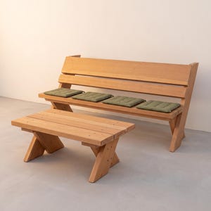 May include: A light brown wooden bench with a high backrest and a matching low coffee table. The bench features four individual olive green rectangular seat cushions. Both pieces of furniture are constructed with angled support legs.