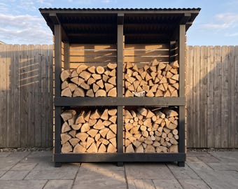 May include: A dark gray wooden firewood storage rack with a corrugated roof, featuring four distinct rectangular compartments filled with stacked, light brown split logs. The structure is open-fronted and has a sturdy frame design.
