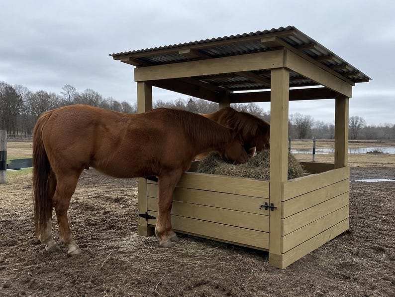 Puede incluir: Un comedero para caballos de madera marr&oacute;n claro con techo de metal corrugado, con un marco cuadrado y paredes de tablones horizontales. Dos caballos alazanes comen heno del interior de la estructura, que tiene bisagras negras.