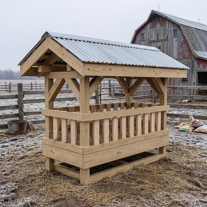 May include: A rectangular wooden hay feeder featuring a gabled roof covered with corrugated metal sheeting. The structure is built with light-colored timber, vertical slats forming the sides, and a sturdy base, designed to hold hay for livestock.