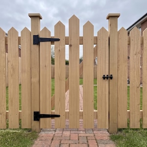 May include: A light brown wooden picket gate featuring pointed tops on each vertical slat. The gate is equipped with two black metal hinges on the left side and a black metal latch on the right side, mounted between two sturdy wooden posts.