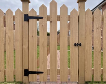 May include: A light brown wooden picket gate featuring pointed tops on each vertical slat. The gate is equipped with two black metal hinges on the left side and a black metal latch on the right side, mounted between two sturdy wooden posts.