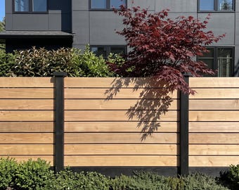 May include: A horizontal wood slat fence featuring light brown planks and dark black vertical support posts. The fence is positioned in front of a dark gray building with multiple windows and is partially obscured by a red Japanese maple tree.