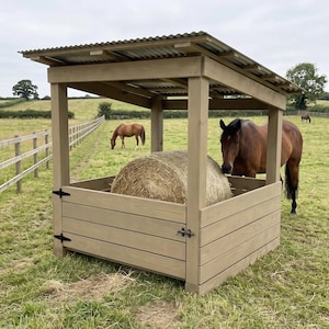 Puede incluir: Un comedero de heno de madera con techo de metal corrugado, con un marco cuadrado y paredes de tablones horizontales. Una gran paca de heno redonda se encuentra dentro, mientras un caballo marr&oacute;n est&aacute; en un campo de hierba.