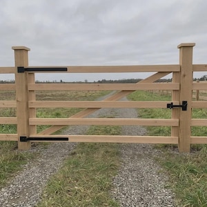 K&ouml;nnte beinhalten: Ein helles Holztor mit vier horizontalen Querlatten und einer diagonalen Verstrebung. Das Tor ist mit schwarzen Metallscharnieren an zwei quadratischen Holzpfosten befestigt und verf&uuml;gt &uuml;ber einen schwarzen Metallriegel auf der rechten Seite.