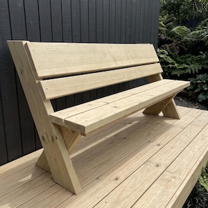 May include: A light-colored wooden bench featuring a backrest composed of two horizontal planks and a seat made of two horizontal planks. The bench has angled side supports that form a V-shape, constructed from natural wood with visible grain.