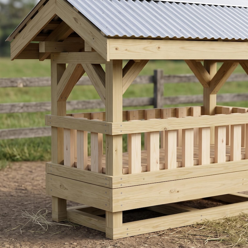 May include: A wooden livestock feeder featuring a rectangular slatted enclosure and a gabled roof covered with corrugated metal sheeting. The structure is built from light-colored timber with visible grain, supported by sturdy corner posts and cross-bracing.