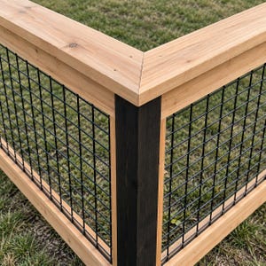 May include: A corner section of a fence featuring natural light-toned wooden horizontal rails and a vertical corner post stained in a dark black finish. The fence panels are filled with a black metal wire grid mesh pattern throughout the structure.