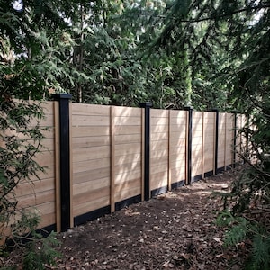 May include: A horizontal wood fence featuring light brown cedar planks arranged in a stacked pattern, supported by vertical black posts and a black base trim, extending across the frame with a natural wood grain texture visible throughout.