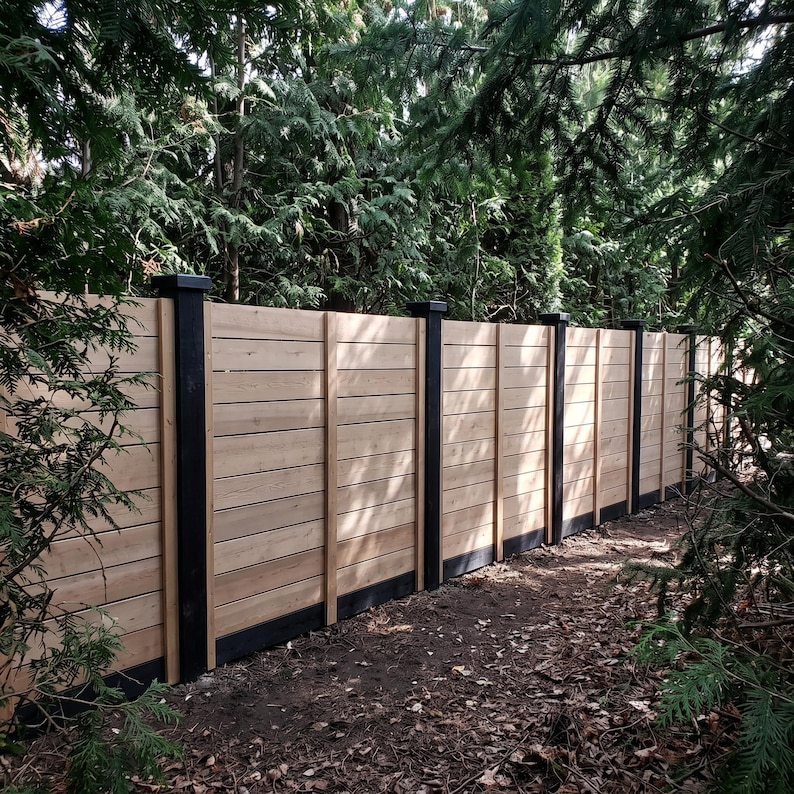 May include: A horizontal wood fence featuring light brown cedar planks arranged in a stacked pattern, supported by vertical black posts and a black base trim, extending across the frame with a natural wood grain texture visible throughout.