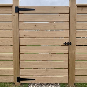 K&ouml;nnte beinhalten: Ein hellbraunes Holztor mit horizontalen Brettern und schwarzen Metallbeschl&auml;gen, bestehend aus zwei gro&szlig;en Langb&auml;ndern auf der linken Seite und einem Schieberiegel auf der rechten Seite, montiert zwischen zwei passenden Holzpfosten.