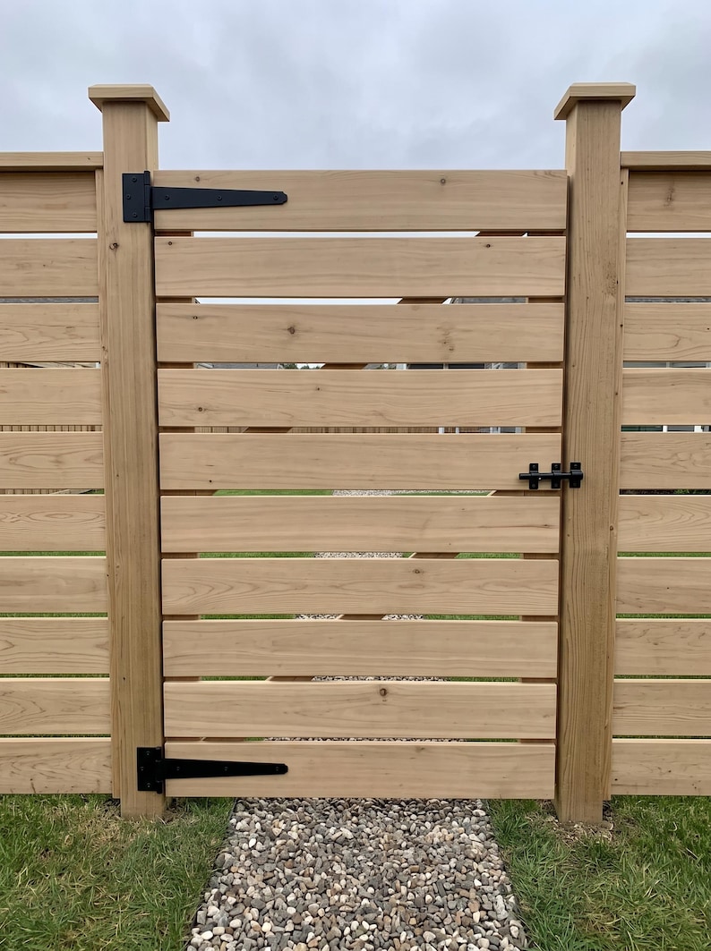 K&ouml;nnte beinhalten: Ein hellbraunes Holztor mit horizontalen Brettern und schwarzen Metallbeschl&auml;gen, bestehend aus zwei gro&szlig;en Langb&auml;ndern auf der linken Seite und einem Schieberiegel auf der rechten Seite, montiert zwischen zwei passenden Holzpfosten.