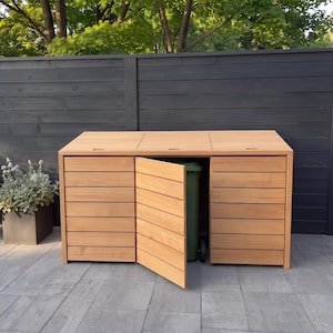 Pode incluir: Um abrigo de madeira castanho-claro para três caixotes do lixo, com ripas horizontais e três tampas articuladas. A porta central está aberta, revelando um caixote do lixo de plástico verde no interior. Estrutura com design retangular.