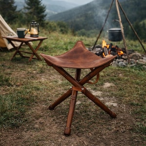 Op de afbeelding: Een bruine leren campingkruk met een driehoekige zitting en houten poten. De kruk staat buiten bij een kampvuur, met een tent en bergen op de achtergrond. De kruk is ontworpen voor gebruik buitenshuis.