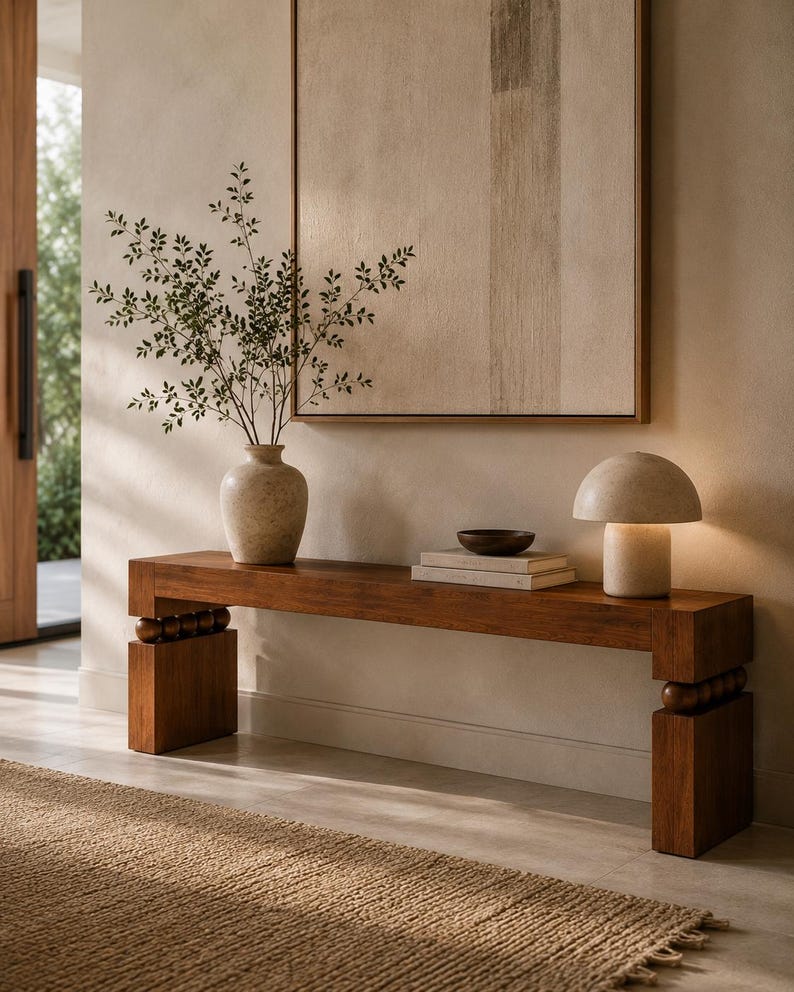May include: A long wooden console table with block legs and decorative wooden spheres, topped with a stone vase containing green branches, two stacked books, a small dark bowl, and a mushroom-shaped stone lamp with a warm light.