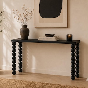 May include: A black console table featuring a flat rectangular top and four legs constructed from stacked spherical wooden balls. The table surface holds a dark brown vase with dried flowers, two stacked books, a small bowl, and a glass candle.