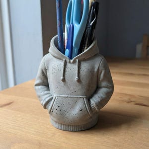 Puede incluir: Un organizador de escritorio de hormigón gris con forma de sudadera con capucha, con un bolsillo delantero y capucha. Contiene tijeras azules, bolígrafos y lápices. La textura moteada le da un aspecto único y moderno.